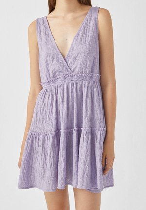 Robe lilas clair avec un décolleté en V profond, sans manches, en tissu texturé et avec un ourlet froncé. Dispose d'une taille froncée pour un ajustement fluide.