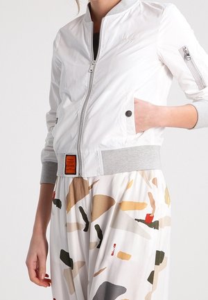 Chaqueta bomber blanca con cierre de cremallera en la parte delantera, puños acanalados grises y un logotipo bordado en el pecho. Combinada con pantalones claros de estampado.