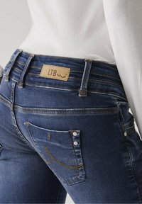 Mörkblå denimjeans med en märkeslogotyp i tan läder, kontraststickningar och dekorativa detaljer på bakfickorna. Fitad design med subtil textur.