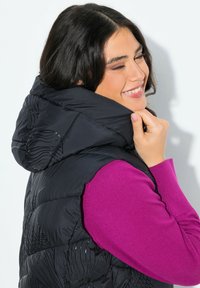 Žena se usmívá se zavřenýma očima, má na sobě černou péřovou vestu přes jasně růžový dlouhý rukáv, přidržuje lem vesty blízko své tváře.