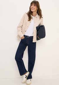 Glunderende vrouw in een beige jasje, witte shirt, donkerblauwe jeans en witte sneakers, leunend tegen de muur, met een opgevouwen marineblauw jasje in haar hand.