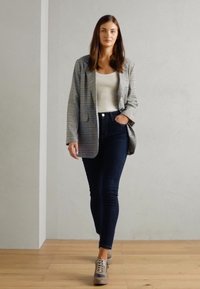 Blazer a cuadros gris y blanco sobre una blusa beige, combinado con jeans oscuros y zapatillas topo. Tela suave, corte relajado y patrones sutiles.