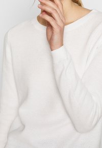 Biały sweter dziany z oddychającą teksturą, z okrągłym dekoltem i ribbami przy mankietach. Zbliżenie na materiał i rękę blisko dekoltu.