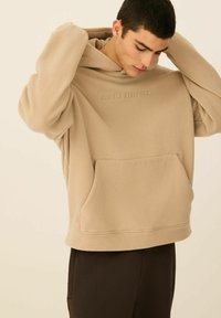 Beżowa bluza z kapturem z przednią kieszenią, wykonana z miękkiego polaru, z wytłoczonym napisem "Diverse Athletics" na piersiach.