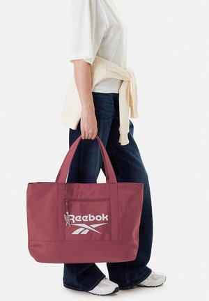 Osoba ubrana w biały top i dżinsy trzyma dużą bordową torbę Reebok z przednią kieszenią na suwak i logo, kremowy sweter zawiązany w pasie.