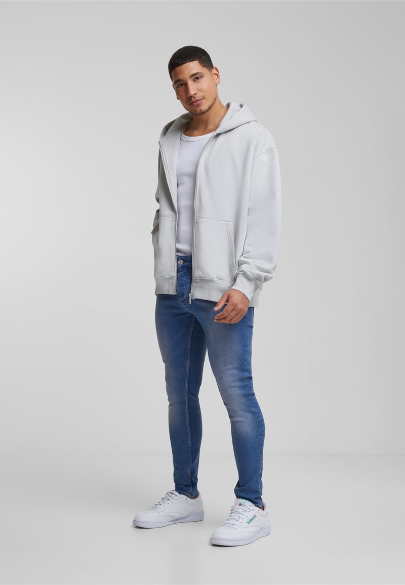 Svijetlosiva zip-up majica s kapuljačom, prednjim džepovima, nosi se preko bijele majice bez rukava i plavih uskih traperica, u paru s bijelim tenisicama.