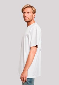 Weißes T-Shirt mit kurzen Ärmeln und Rundhalsausschnitt, mit einem Druckdesign auf der Rückseite. Das Modell trägt hellblaue beschichtete Jeans.