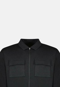 Zwarte jas met een volledige ritssluiting, puntige kraag en twee borstzakken. Gemaakt van een gladdere stof met een matte afwerking.