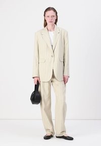 Completo a righe beige realizzato in tessuto leggero, con blazer a un bottone e pantaloni a gamba dritta. Il modello tiene in mano una piccola borsa nera.
