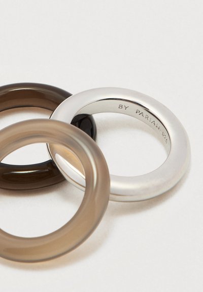 Trois anneaux disposés ensemble : un en métal argenté, un translucide marron, et un noir, présentant des textures lisses et des designs simples et arrondis.