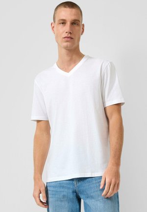 Młody mężczyzna z krótkimi włosami, ubrany w gładką białą koszulkę z dekoltem w serek oraz niebieskie dżinsy na jednolitym szarym tle.