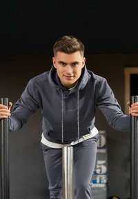 Grijze hoodie met koordjes, een voorzak en bijpassende grijze broek. Het model leunt op twee zwarte stokken, tegen een donkere achtergrond.