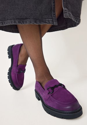 Mocassins en suede violet avec une semelle épaisse noire et un détail de quincaillerie décoratif. Texture lisse avec un embout arrondi et un petit talon.
