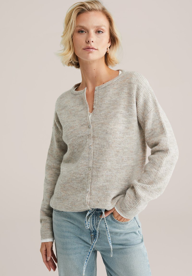 Cardigan gris clair avec une texture côtelée, doté d'un col rond, de boutons à l'avant et de manches longues. Associé à un jean bleu clair.
