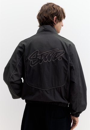 Giovane uomo con capelli castani corti che indossa una giacca nera larga con una scritta ricamata in rilievo sulla schiena, su uno sfondo chiaro e uniforme.