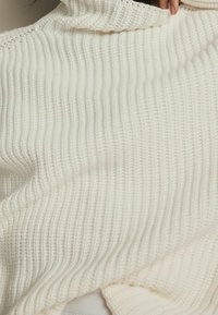 Cremefarbener Strickpullover mit gerippter Struktur, der einen hohen Kragen und einen lockeren Schnitt aufweist, was sein weiches und gemütliches Aussehen unterstreicht.