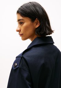 Cappotto trench blu navy con colletto strutturato, grande dettaglio con bottoni sulla spalla e texture liscia, mostrato da un profilo laterale.