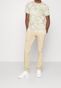 Světlé béžové camo tričko s přední kapsou, zkombinované s khaki chinos kalhotami a bílými teniskami. Měkká tkanina s uvolněným střihem a hladkou texturou.