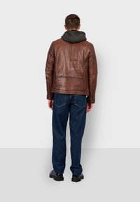 Bruine leren jas met capuchon, gekenmerkt door een gladde textuur en een ontspannen pasvorm, gecombineerd met donkerblauwe denim broek en zwarte schoenen.