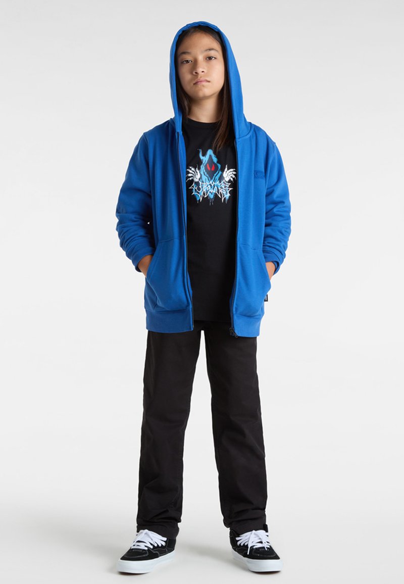 Sudadera azul con cremallera y capucha, camiseta gráfica negra con diseños intrincados, pantalones negros y zapatillas negras con cordones blancos.
