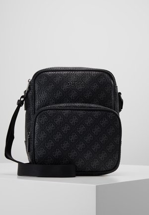 Sac bandoulière Guess noir avec motif de logo discret sur toute la surface, poche avant zippée et sangle ajustable, posé sur une surface blanche.