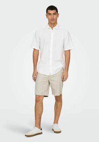 White short-sleeved button-up shirt, beige shorts, white sneakers with brown accents. Model stands against a plain light background.