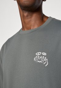 T-shirt cinza de algodão com decote redondo, apresentando um design gráfico branco com o texto "KARMA" e ilustrações de mãos na área do lado esquerdo do peito.