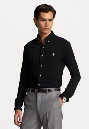 Camicia nera a maniche lunghe con colletto button-down, logo bianco sul petto, abbinata a pantaloni grigi su misura e cintura in pelle nera.