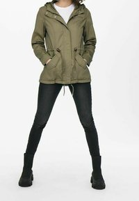 Olivgrön huoodjacka med dragkedja fram, dragsko i midjan och patchfickor. Bärs över en vit skjorta och svarta slim-fit jeans, med svarta stövlar.