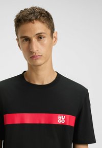 T-shirt in cotone nero con una vistosa striscia rossa orizzontale e il logo bianco "HUGO". Maniche corte e design con collo rotondo.