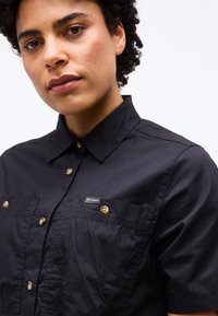 Νεαρό άτομο με σγουρά μαλλιά που φοράει μαύρο πουκάμισο με κουμπιά και τσέπες στο στήθος, με ένα μικρό λογότυπο Columbia στην τσέπη.