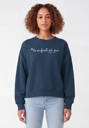 Vrouw met krullend haar die een donkerblauwe trui draagt met de tekst "So einfach ist das." en lichtblauwe spijkerbroek, staand tegen een effen achtergrond.