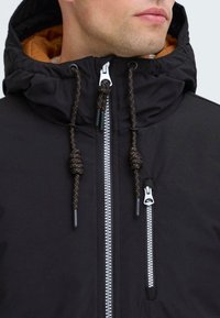 Man wearing black hooded jacket with orange lining, featuring two silver zippers and black drawstrings with orange accents.