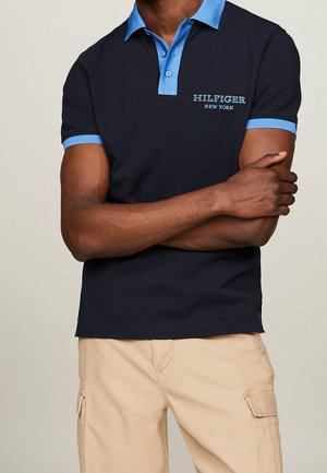 Man wearing een marineblauw poloshirt met lichtblauwe kraag en mouwranden, met het logo "Hilfiger New York", en beige cargobroek, armen gekruist.
