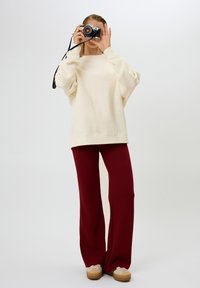 Felpa oversize color crema con spalle basse, abbinata a pantaloni larghi bordeaux e sneakers beige. La persona tiene in mano una macchina fotografica vintage.