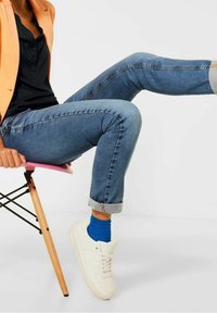 Jasnoniebieskie dżinsy z podwiniętymi mankietami, w zestawieniu z białymi trampkami i niebieskimi skarpetkami. Czarna koszula i pomarańczowy żakiet dopełniają stylizację.