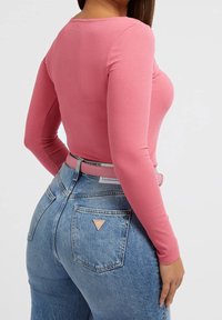 Donna che indossa un top lungo aderente a coste rosa infilato in jeans blu di denim a vita alta con una cintura rosa, vista da dietro.