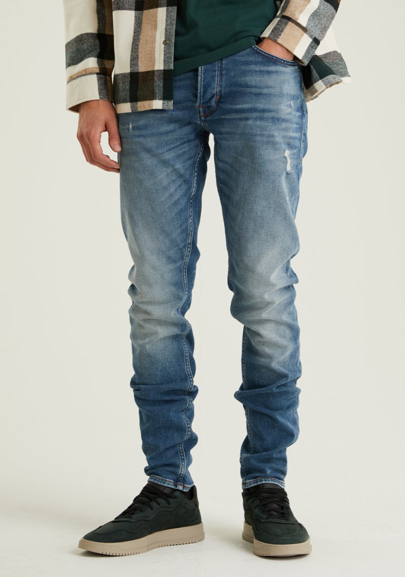 Világoskék, slim fit farmernadrág halvány mosással és enyhe kopással; sötétzöld sportcipővel és kockás flanelinggel párosítva.