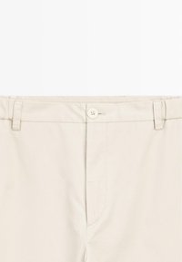 Taille de pantalon beige avec fermeture par bouton et passants pour ceinture, montrant les détails des coutures avant.