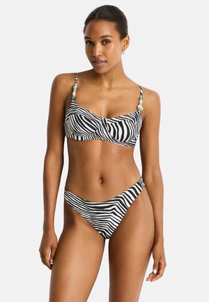 Kobieta w czarno-białym bikini w panterkę z koralikowymi ramiączkami, stojąca na jednolitym jasnym tle.