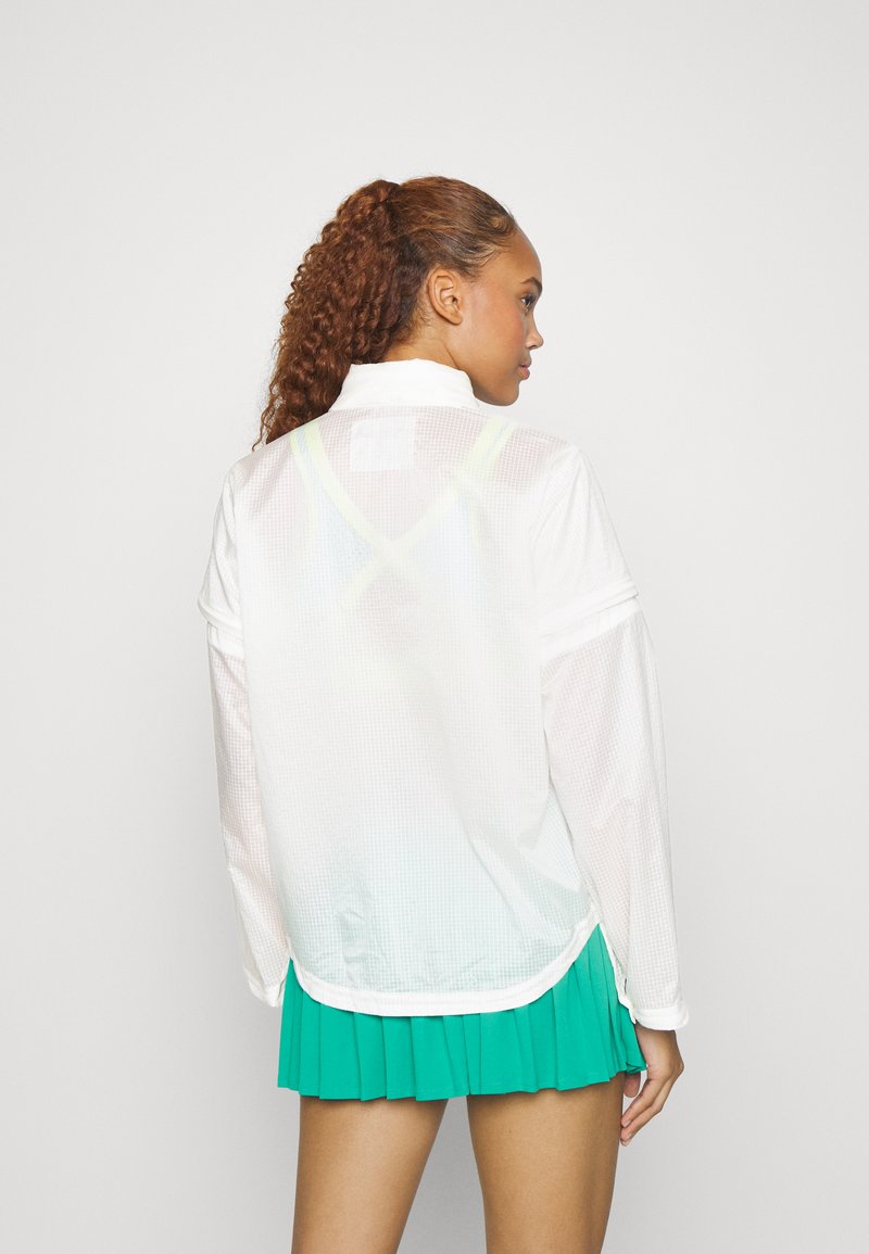 Femme aux cheveux bouclés portant un haut blanc en voile à manches longues, laissant apparaître un soutien-gorge de sport en dessous, associé à une jupe plissée verte, de dos.