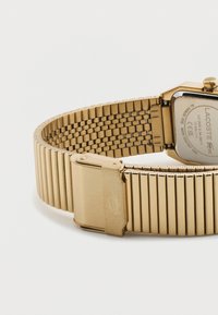 Χρυστό μεταλλικό λουράκι ρολογιού με κούμπωμα που εμφανίζει το χαραγμένο λογότυπο κροκόδειλου, συνδεδεμένο με ορθογώνια πίσω όψη θήκης ρολογιού χαραγμένη με λογότυπο και λεπτομέρειες αντοχής στο νερό.