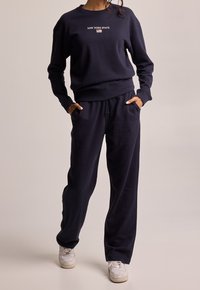 Námořnický modrý svetr s textem "NEW YORK STATE" a malým detailem vlajky, doplněný volně střiženými tepláky a bílými teniskami.