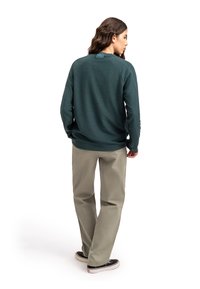 Maglietta verde scuro a maniche lunghe con maniche raglan, dotata di un etichetta sul retro. Abbinata a pantaloni beige chiaro e scarpe nere.