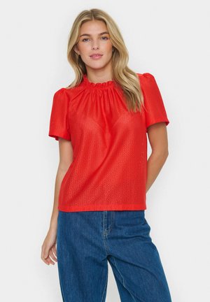Rotes Kurzarmoberteil mit gerafftem Ausschnitt, strukturierter Rautenoptik und lässiger Passform. Kombiniert mit Jeans.