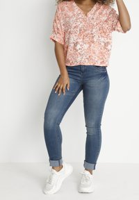 Blusa a fiori con bottoni color pesca e bianco, abbinata a jeans skinny blu. Il modello indossa sneakers bianche. Tessuto morbido e taglio rilassato.