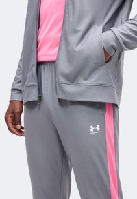 Άνδρας φοράει γκρι φόρμα Under Armour με ροζ πλευρικές ρίγες και ροζ πουκάμισο, χέρι στην τσέπη του μπουφάν.