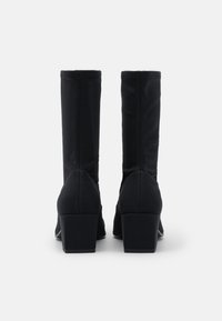 Botas de tacón negro con un upper de tela elástica y ajustada. Tacones cuadrados y textura suave. Diseño simple sin hardware o adornos visibles.