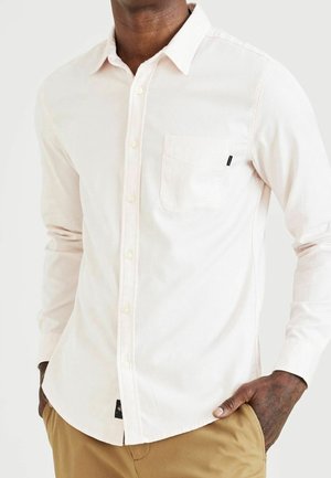 Homme portant une chemise blanche à manches longues avec poche poitrine et un pantalon beige, les mains dans les poches, debout devant un fond uni.