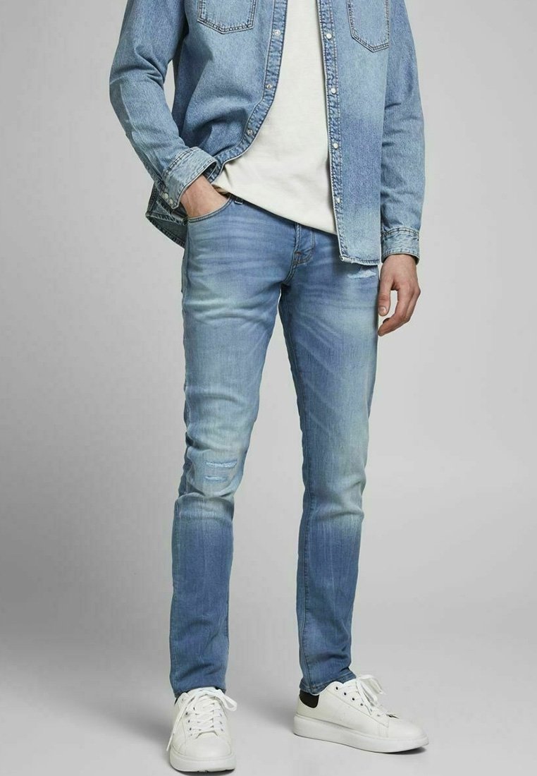Homme portant un jean et une veste en denim bleu clair, un t-shirt blanc, les mains dans les poches, et des baskets blanches sur un fond gris.
